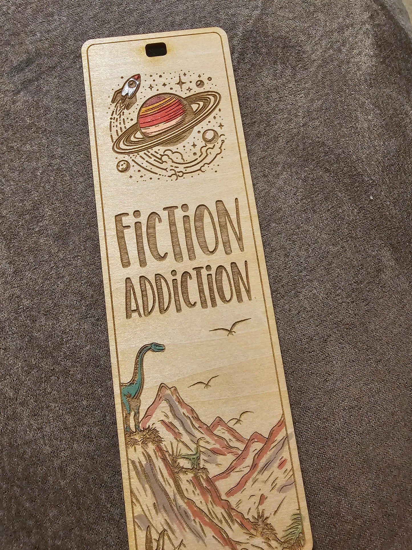 Wooden engraved bookmark with 'Fiction Addiction', Saturn, rocket, dinosaur, and mountain art