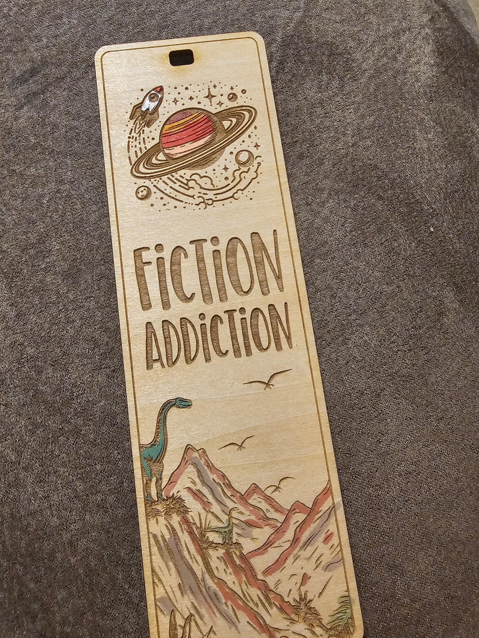 Wooden engraved bookmark with 'Fiction Addiction', Saturn, rocket, dinosaur, and mountain art