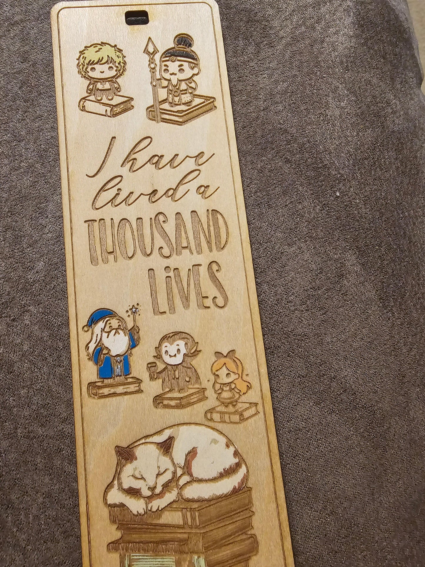 Custom wooden bookmark engraved with cute fantasy characters, books, a cat, and the phrase 'I have lived a thousand lives', from BearNugget Customs.